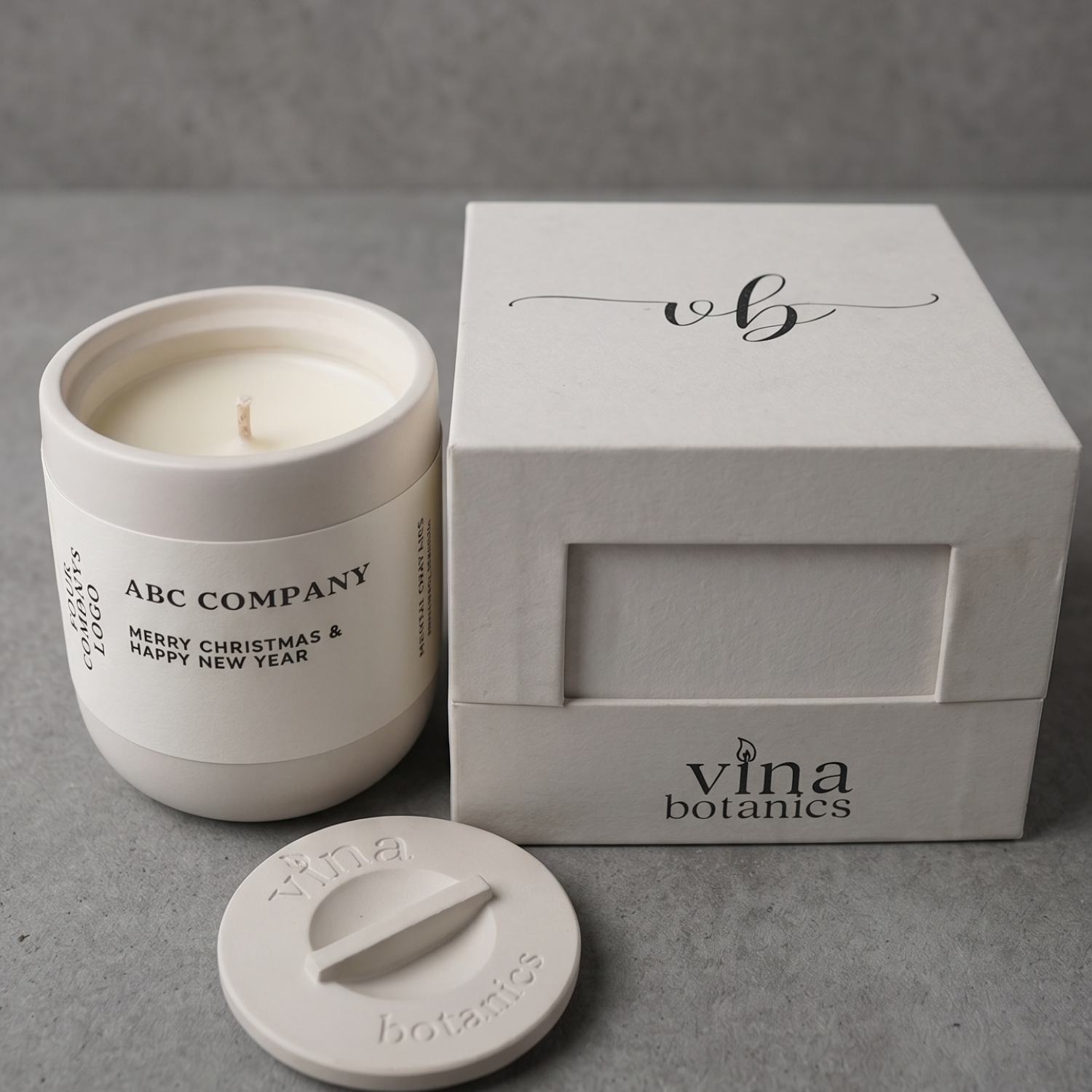 White candle with lid and 'ABC Company' branding next to a 'vina botanics' branded box on a gray surface.