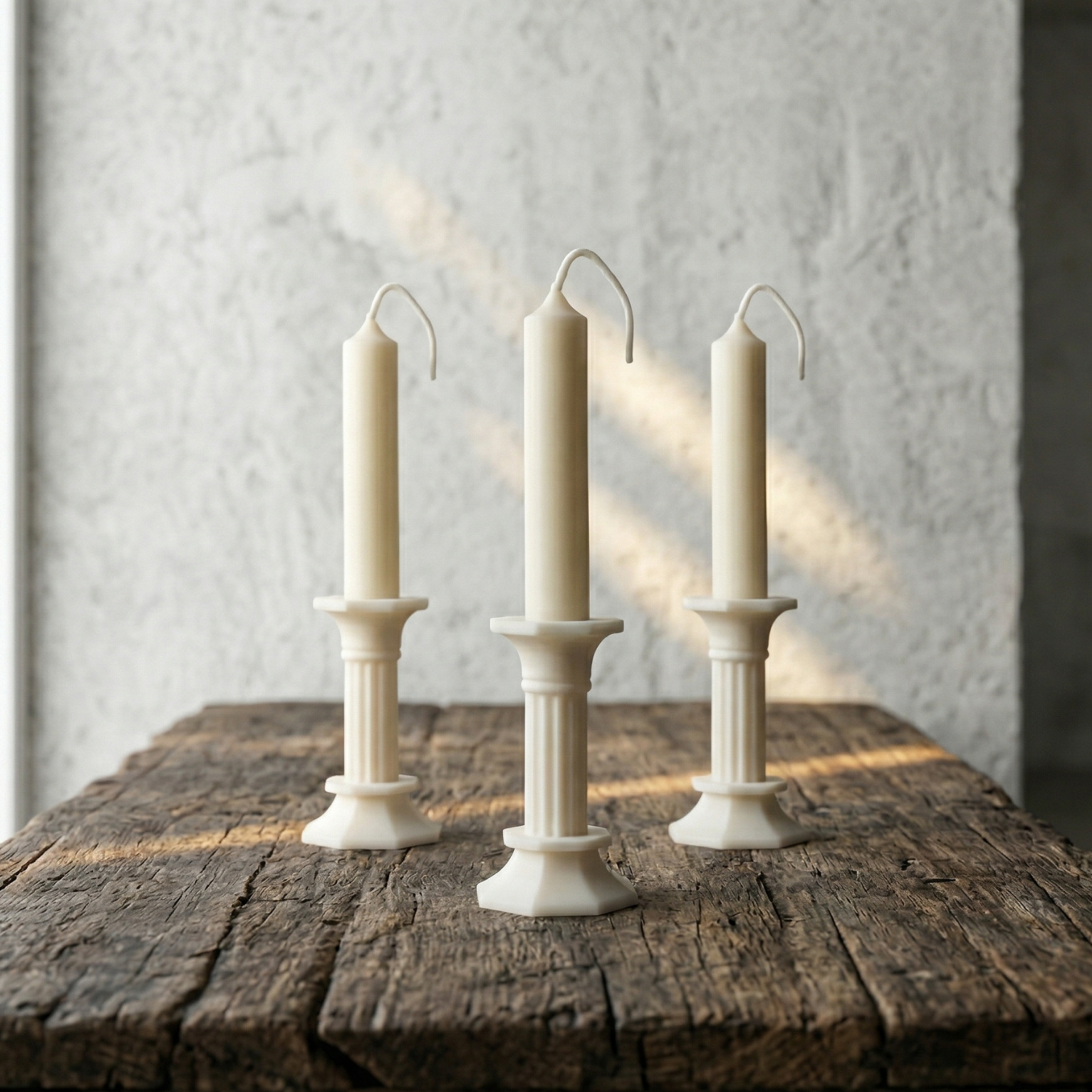 Three white candles in decorative candlesticks on a wooden surface with a textured wall background.